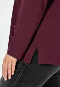Bordeaux langlijfshirt met een relaxte fit, voorzien van zijsplitten en een zachte textuur. Gecombineerd met donkere denim jeans.