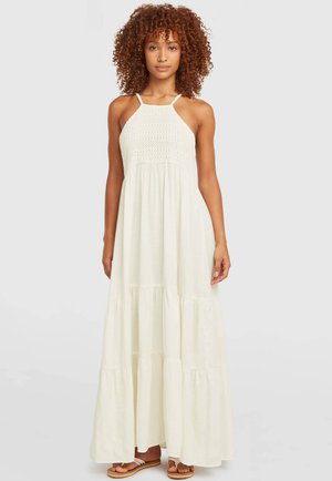 Ženska s kodrastimi lasmi, oblečena v kremasto maxi obleko brez rokavov z zgornjim delom na kvačkanje in večplastno krilo, stoji pred enobarvnim ozadjem.
