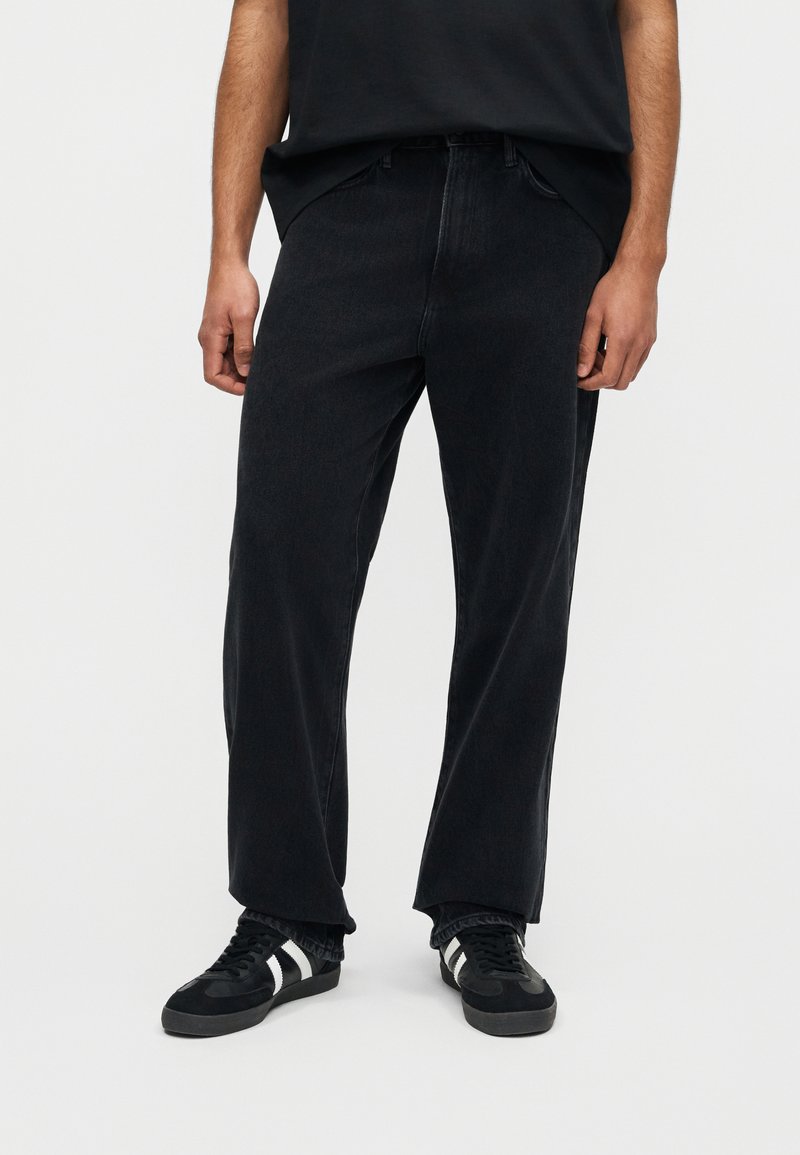 Mand iført løstsiddende sorte jeans, sorte sneakers med hvide striber og en sort skjorte, stående foran en ensfarvet hvid baggrund.