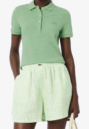 Vrouw draagt een lichtgroen poloshirt met korte mouwen en een wit-groen gestreepte short met hoge taille, en houdt een beige tas vast.