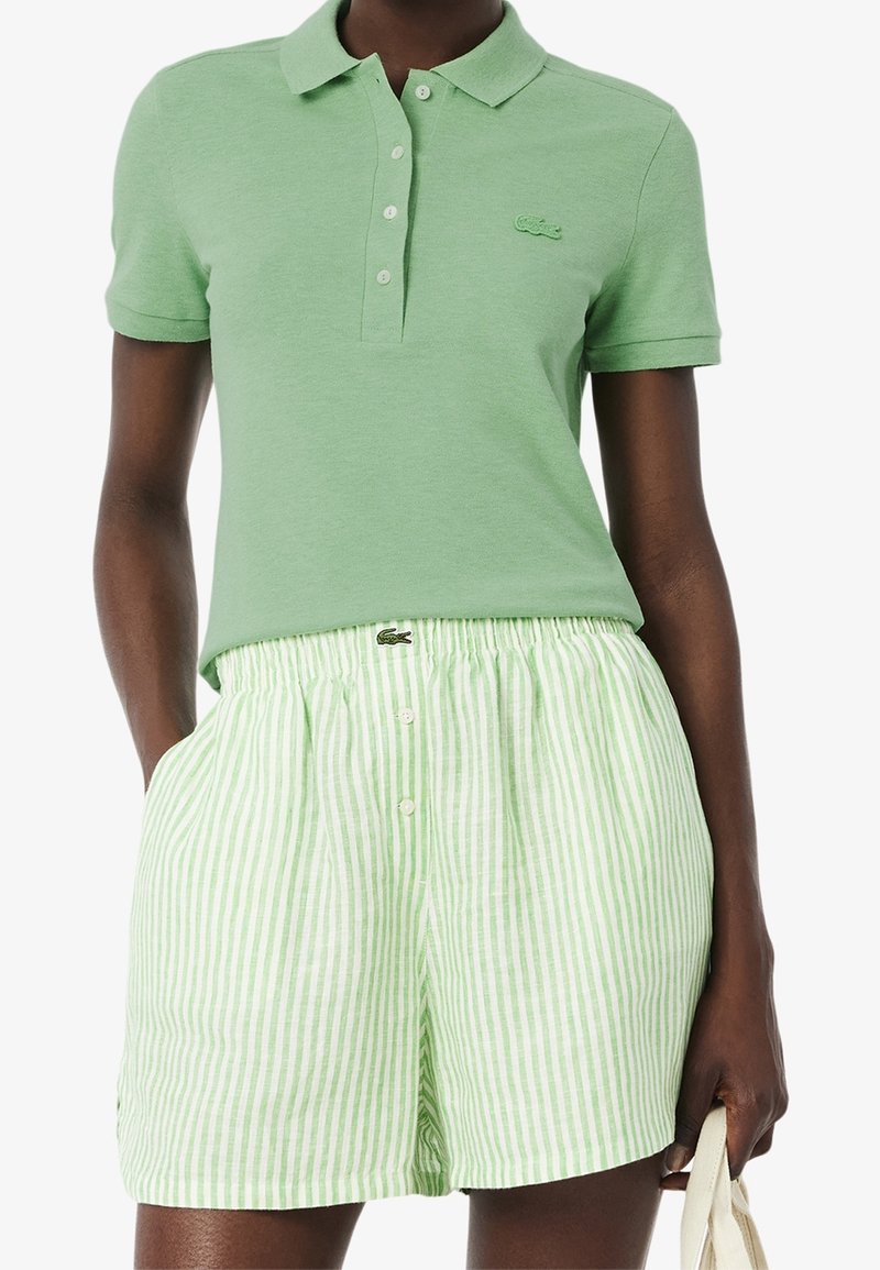 Femme portant un polo à manches courtes vert clair et un short taille haute rayé blanc et vert, tenant un sac beige.