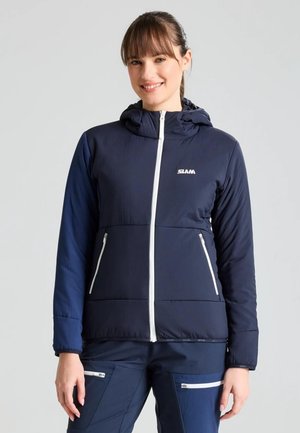 Una mujer sonriente luce una chaqueta azul marino con capucha y cremalleras blancas, junto con unos pantalones a juego, sobre un fondo claro y sencillo.