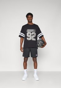 Svart oversized t-skjorte med "CHAMPION 92" trykk, hvite striper på ermene, svarte shorts, hvite sneakers, holder en basketball.