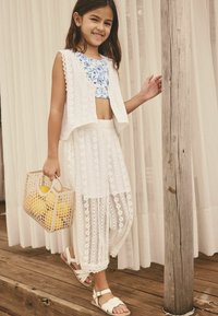 White crocheted vest and wide-leg pants with floral blue crop top. Holding beige woven bag with yellow items. Sandals with strap detailing.