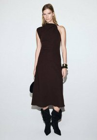 Vestido marrón sin mangas con un corsé ajustado y una silueta holgada, combinado con botas negras de caña alta y un bolso pequeño.