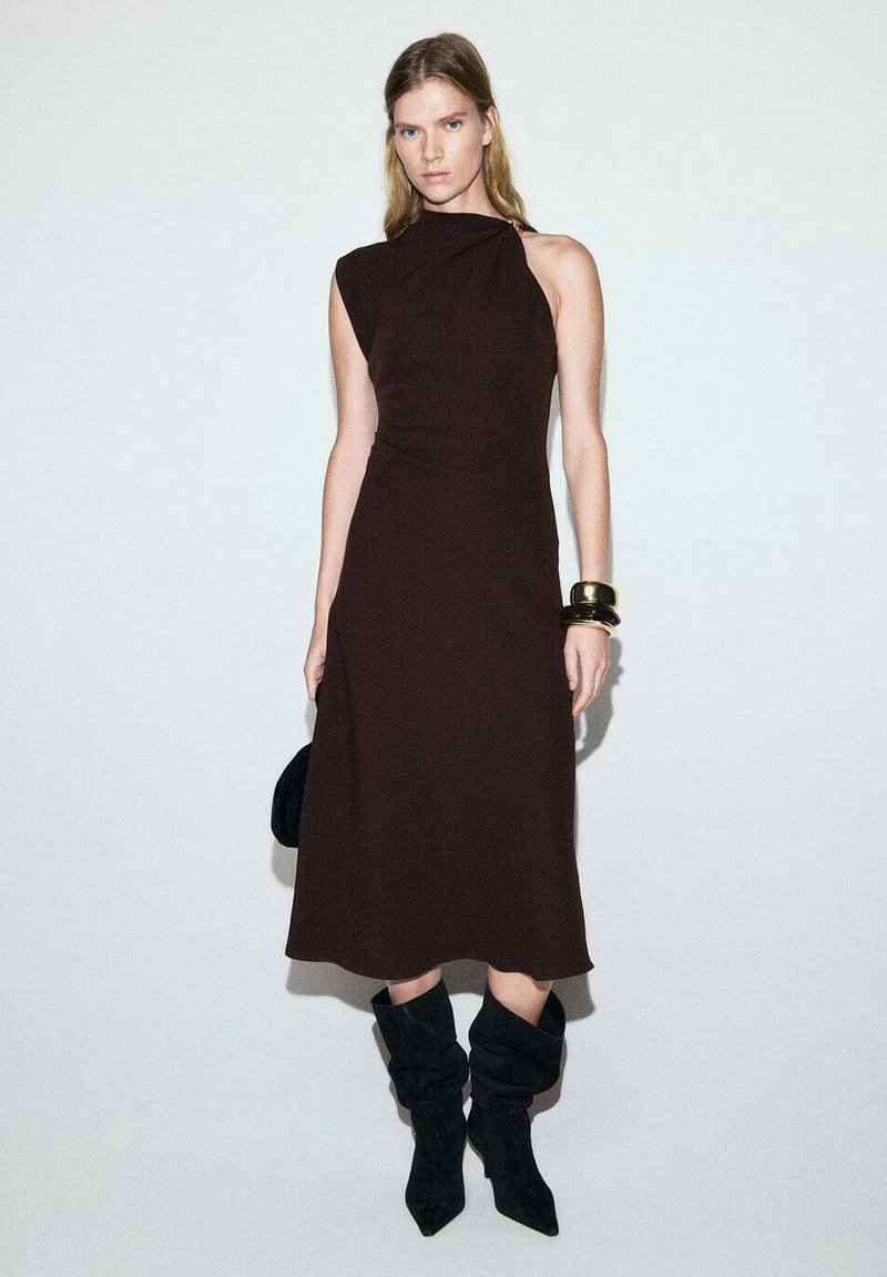 Vestido marrón sin mangas con un corsé ajustado y una silueta holgada, combinado con botas negras de caña alta y un bolso pequeño.