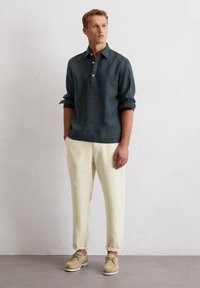 Chemise en lin vert foncé avec un col boutonné et une poche sur la poitrine, associée à un pantalon en lin crème et des chaussures à lacets beiges.