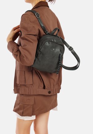 Zaino in pelle nera con spallacci regolabili, forma rotonda, cerniera laterale e superficie testurizzata, indossato sopra una giacca marrone.