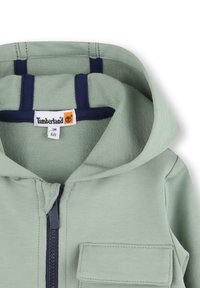 Lichtgroene rits-hoodie met een marineblauwe accent in de capuchon. Heeft een voorzak en een zachte, gestructureerde stof. Timberland-label zichtbaar.