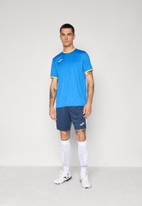 Blue athletic shirt with yellow trim, paired with navy shorts featuring white accents. White knee-high socks with grip dots; white sports shoes.
