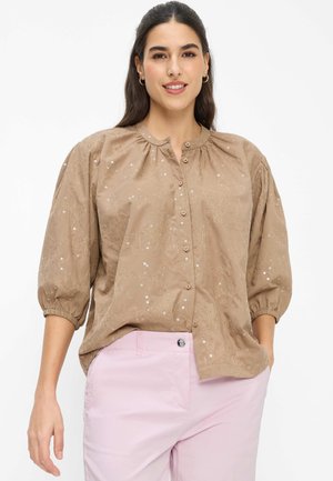 Frau trägt eine hellbraune bestickte Bluse mit Knöpfen, drei Viertel Puffärmel und hellrosa Hose, lächelt mit einer Hand in der Tasche.