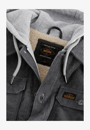 Giacca in velluto grigio con cappuccio grigio, foderata in pile e con etichetta nera. Presenta una zip, bottoni a pressione e tessuto testurizzato.