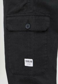 Black cargo pants with a patch pocket, featuring a button closure and a white brand label stitching. Fabric has a smooth texture.