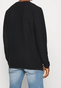 Black textured long-sleeve shirt with a ribbed design, round neckline, and a relaxed fit, paired with light blue jeans.