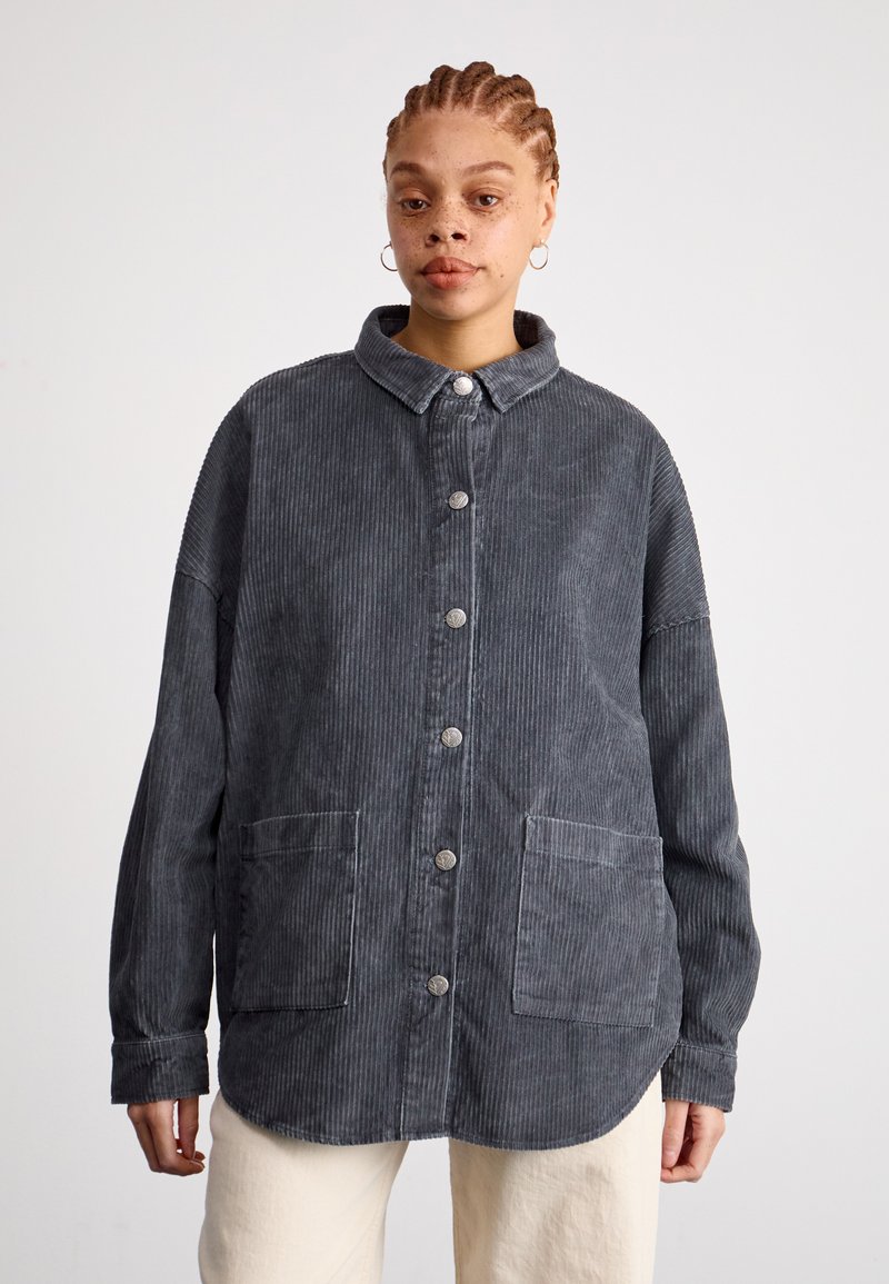 Sivá korduroy bunda s golierom, predným zapínaním na gombíky, dvoma veľkými vreckami a dlhými rukávmi; s vertikálnou ryhovanou textúrou.