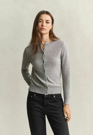Jonge vrouw met lang bruin haar, gekleed in een grijze gebuttonede cardigan en een zwarte spijkerbroek, staand tegen een eenvoudige lichte achtergrond.