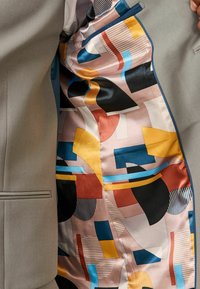 Chaqueta de traje gris con un forro de seda estampada que presenta formas abstractas en azul, naranja, rojo y negro sobre un fondo claro.