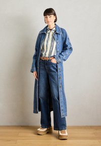 Lange denimjas met knopen aan de voorkant, gecombineerd met een gestreept shirt en wijde jeans. Beige plateau schoenen met bontaccenten. Licht houten vloer.
