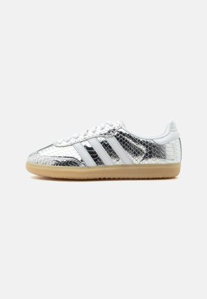 Lavtop sneaker med sølv slangeskind tekstur, hvide striber, hvide snørebånd og lys brun gummisål på en ensfarvet baggrund.
