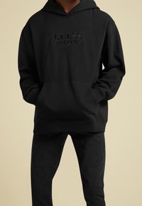 Persona con una felpa nera con "GUESS ORIGINALS" impresso sul petto, abbinata a jeans neri, mani nelle tasche della felpa.