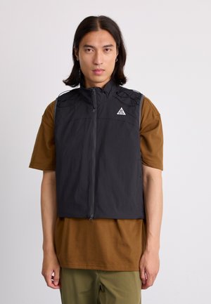 Black sleeveless vest with high collar, zip front, and exterior cord details. Worn over a brown shirt, paired with green pants.