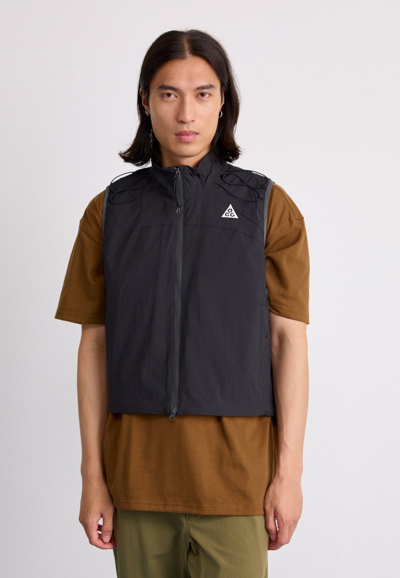 Black sleeveless vest with high collar, zip front, and exterior cord details. Worn over a brown shirt, paired with green pants.