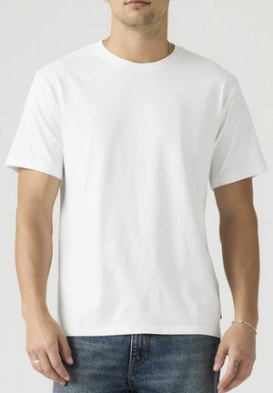 Homme portant un t-shirt blanc uni à manches courtes et col rond, ainsi qu'un jean bleu, debout avec les bras détendus le long du corps sur un fond neutre.