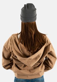 Vrouw met lang bruin haar, gekleed in een grijze beanie, een tan hoodie en blauwe spijkerbroek, die met de rug naar de camera staat tegen een eenvoudige lichte achtergrond.