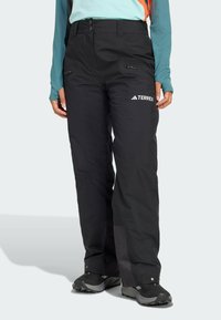 Čierne nepremokavé vonkajšie nohavice so zipsovými vreckami, strihom do tvaru a spevnenými manžetami. Obsahujú logo na stehne a zapínanie na gombík.