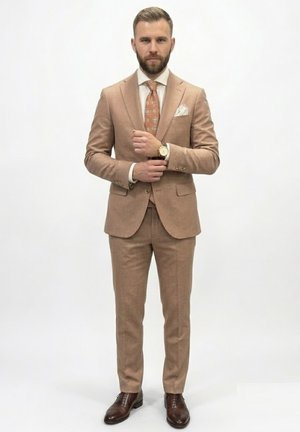 Hombre con traje beige ajustando el gemelo, lleva corbata naranja con estampado, camisa blanca, pañuelo en el bolsillo y zapatos marrones de vestir, de pie sobre fondo blanco.