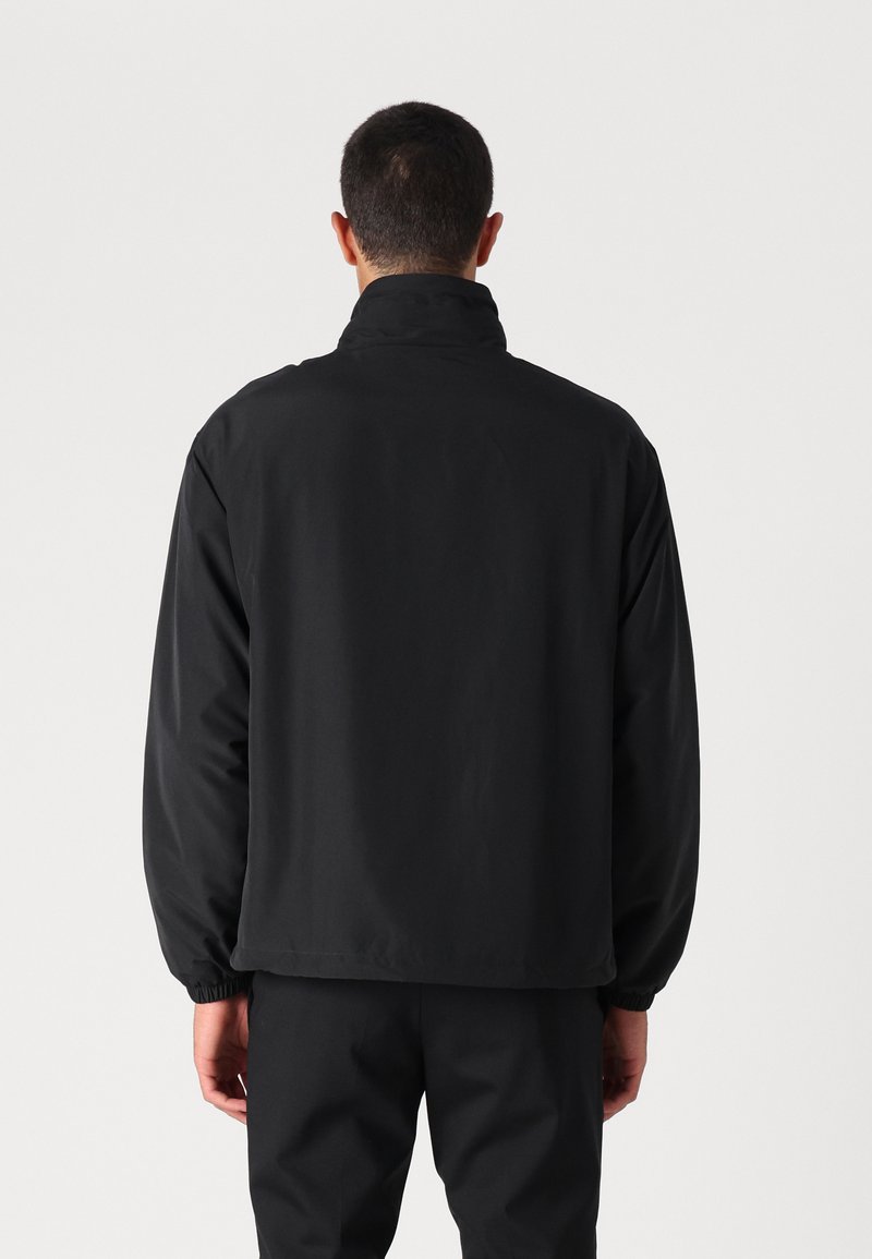Veste noire légère avec col montant et poignets élastiques. Présente une texture lisse et mate ainsi qu'un design à coupe décontractée.