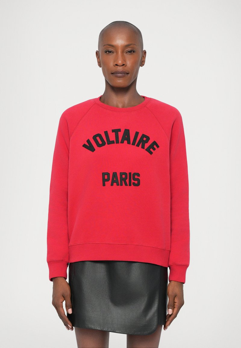 Rode sweatshirt met de tekst "VOLTAIRE PARIS" in zwart over de voorkant. Lange mouwen, ronde hals, gecombineerd met een zwarte faux leren rok.