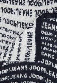 Gebreide sjaal met een patroon van gedurfde, herhalende tekst in marineblauw op een crèmekleurige achtergrond, met een zachte, gestructureerde afwerking.