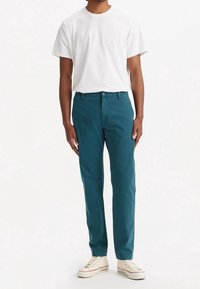 Pantaloni di cotone turchese con taglio dritto, abbinati a una t-shirt bianca semplice e sneakers beige con accenti rossi. Design semplice, dettagli minimi.