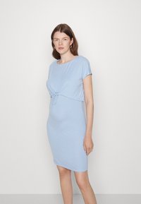 Vestido de maternidade azul claro em tecido suave, com mangas curtas, detalhe frontal em nó e silhueta ajustada. Textura lisa por todo o vestido.