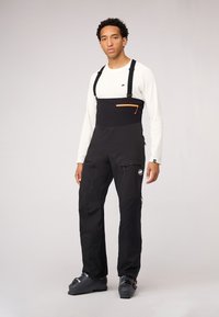 Schwarze Schneehosen mit verstellbaren Trägern, orangefarbener Reißverschlusstasche, Cargo-Taschen und einem Logodetail. Getragen über einem weißen Langarmshirt.