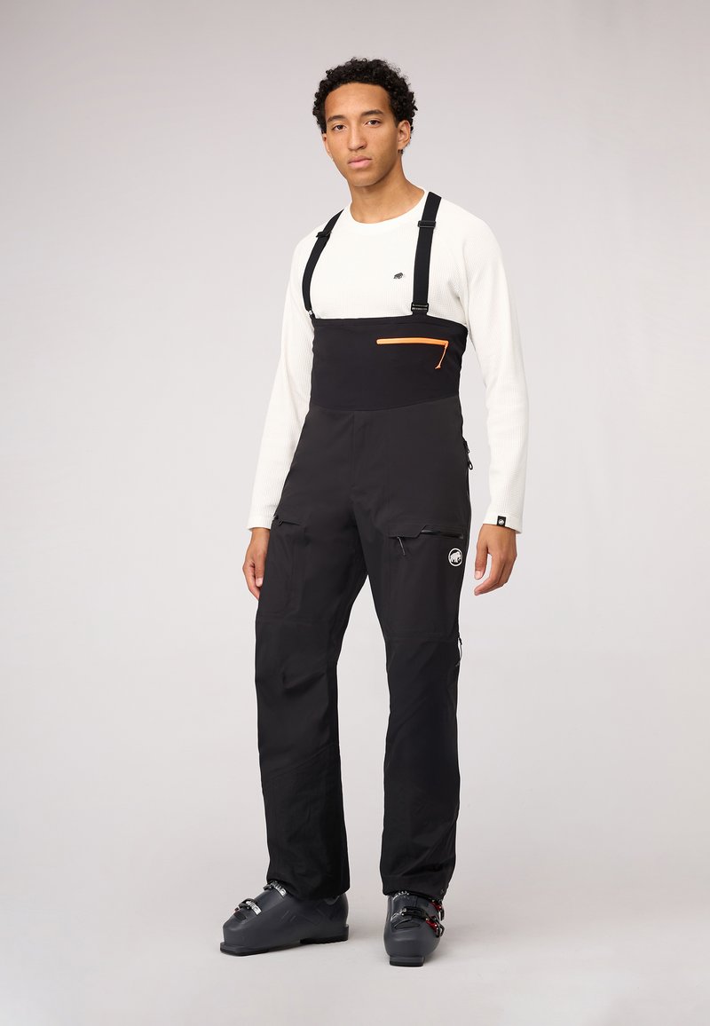 Schwarze Schneehosen mit verstellbaren Trägern, orangefarbener Reißverschlusstasche, Cargo-Taschen und einem Logodetail. Getragen über einem weißen Langarmshirt.
