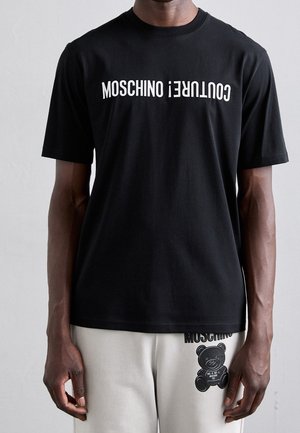 Czarny bawełniany t-shirt z wyraźnym białym napisem "MOSCHINO" i "COUNTER." Okrągły dekolt i krótkie rękawy, w połączeniu z jasnymi spodniami.