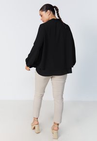 Black, loose-fitting blouse with long sleeves and ruffled neckline, paired with beige fitted pants and wedge-heeled sandals, viewed from behind.