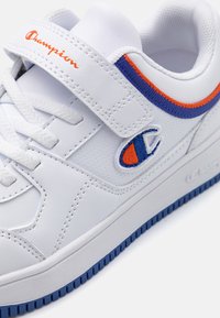 Sneaker bianco con strappo in velcro, tomaia in sintetico testurizzato, con accenti blu e arancioni sui lati e suola in gomma testurizzata.