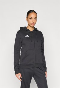 Μαύρο φούτερ με φερμουάρ από μαλακό υλικό, που διαθέτει κουκούλα, μπροστινές τσέπες και λευκό λογότυπο Adidas στη ν πάνω αριστερή πλευρά.