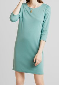 Ljust teal, knälång klänning i mjukt, stretcht tyg. Har en rund halsringning och trekvartslånga ärmar. Minimalistisk design utan mönster.