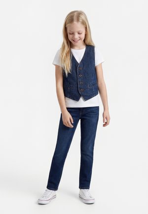 Noor tüdruk seisab, kandmas valget t-särki, nööbitud tumedat teksaliivi, tumedaid teksapükse ja valgeid tossusid, vaadates allapoole naeratades.