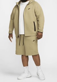 Kaki Nike sweatshirts med dragkedja, matchande shorts och vit t-shirt. Har en logotyp, dragsnören och fickor med dragkedja. Vita sneakers ingår.