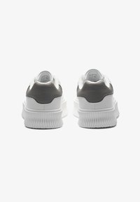 Hvite sneakers med en teksturert grå bakdel, tykk hvit såle og avrundet form. Har fremtredende riller på yttersålen.
