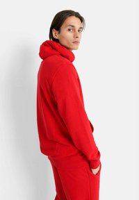 Ung man som står på sidan i en röd hoodie och matchande röda byxor med ena handen i fickan mot en vit bakgrund.