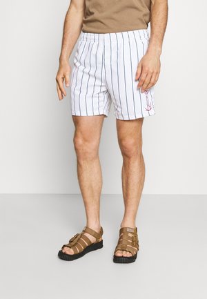 Mann trägt weiße Shorts mit vertikalen blauen Streifen und einem kleinen roten Anker auf dem linken Bein, kombiniert mit braunen Sandalen.