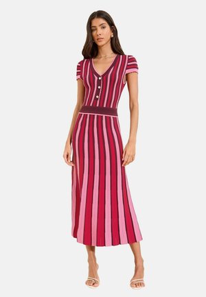 Femeie purtând o rochie midi cu mâneci scurte, cu nasturi, decolteu în V, cu dungi verticale roz, roșii și bordo, asortată cu sandale albe cu degetele descoperite.