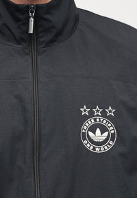 Chaqueta negra con cremallera y cuello alto, con un logo blanco bordado y tres estrellas encima. Textura suave con un brillo sutil.