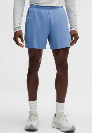 Mann trägt hellblaue Sportshorts, weißes Langarmshirt, weiße Socken und weiße Laufschuhe, steht mit entspannten Armen.